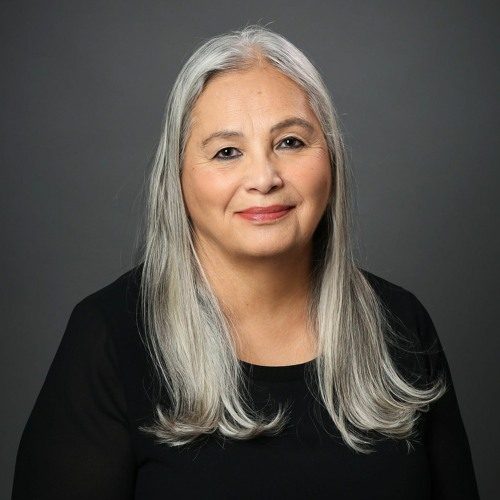 Headshot of Denise Lajimodiere. She is wearing a black shirt and her long gray hair is down around her shoulders as she smiles softly.