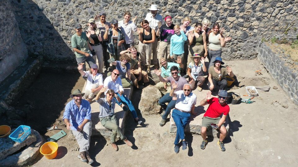 Large group of people wave up at the camera from an archaeological trench in Pompeii.