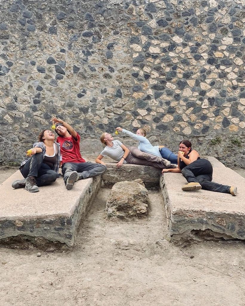 Archaeology students in Pompeii feed each other fruit as if they are parodying an old painting.