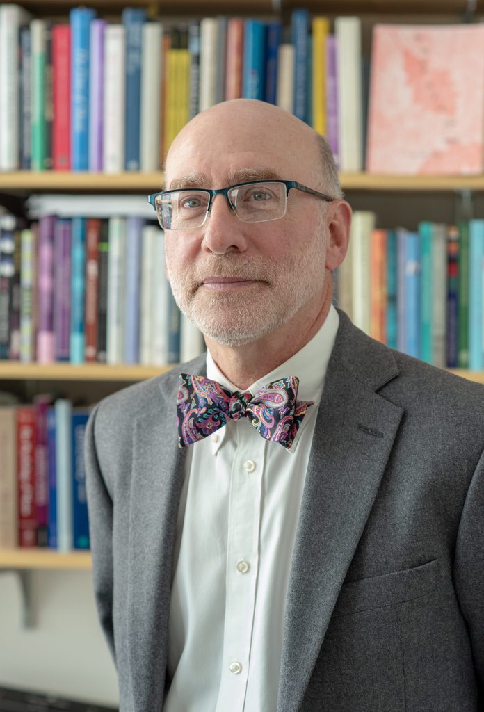 Justin London in a suit and colorful bowtie in front of full bookshelves.