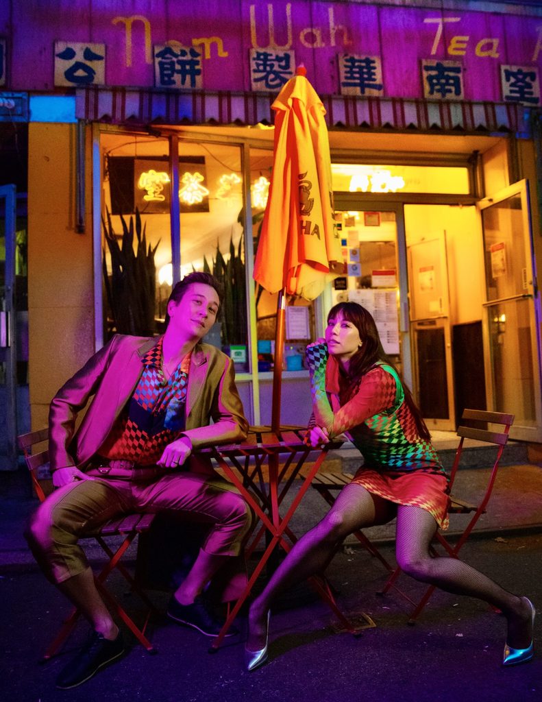 Phil Chan and Georgina Pazcoguin sit artfully in colorful outfits underneath colorful lights in front of a tea shop in Chinatown.