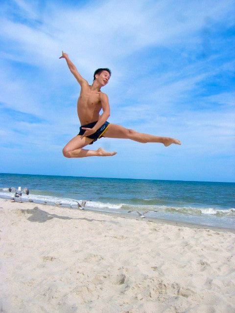Phil Chan leaps high into the air and strikes a ballet pose on a beach.