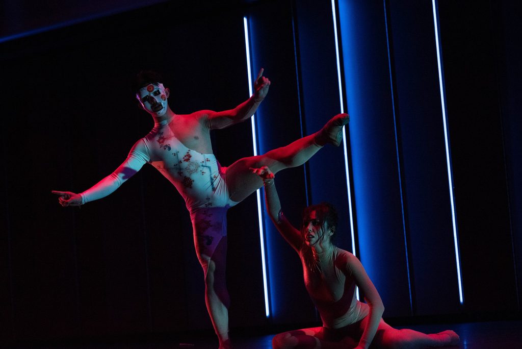 A ballet dancer balances on one foot while spreading the rest of his limbs out wide. Another dancer is collapsed near him with a distressed expression. They are highlighted in red light while blue lights them from behind.