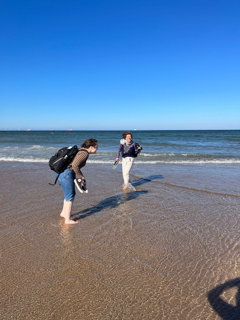 Margaret De Fer ’24 and Serena Zabin wade into waves on the beach, holding up their shoes in their hands.