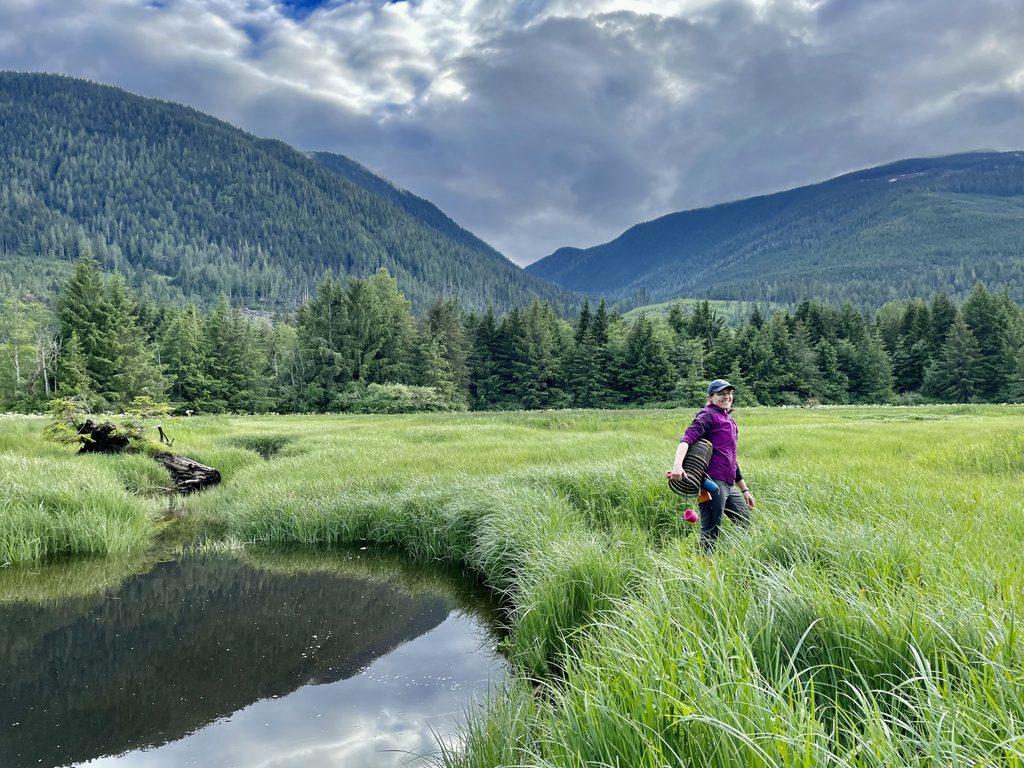 Amanda Hund, carrying a basket, smiles at the camera next to water and in front of large, tree-covered mountains.