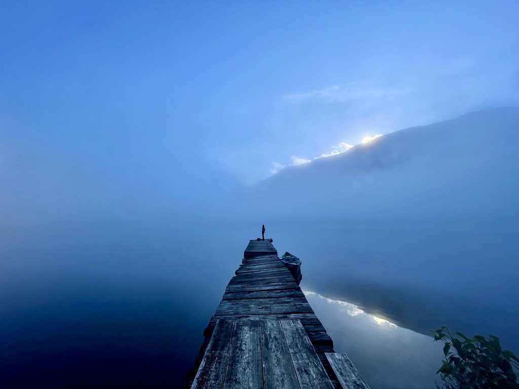 View through heavy mist of a person at the end of a dock with a canoe floating beside it as the sun barely peeks over distant mountains.