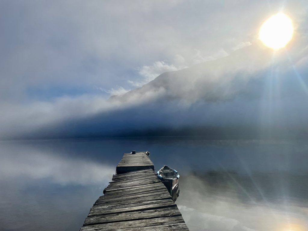 View through the mist of an empty dock with a canoe floating beside it as the sun rises over distant mountains.