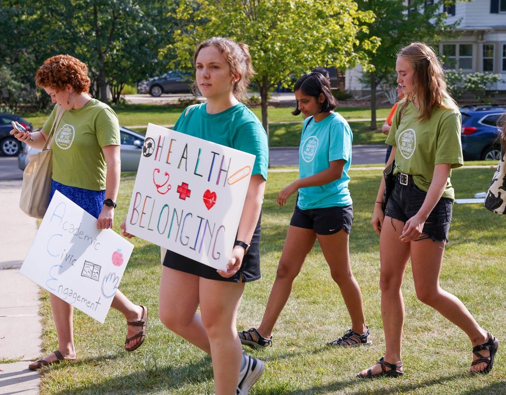 A student in a CCCE shirt carries a poster reading "Health and Belonging." Behind them are more students in CCCE shirts, with one of them carrying a poster reading "Academic Civic Engagement."