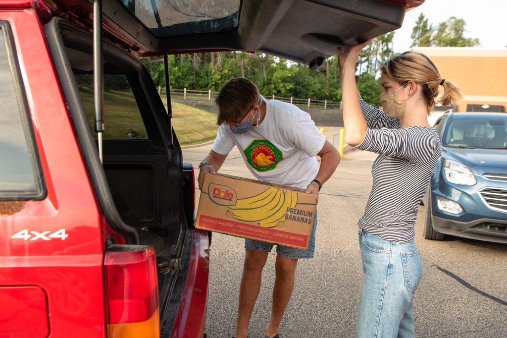 Two students load a cardboard box of food into the back of a car.