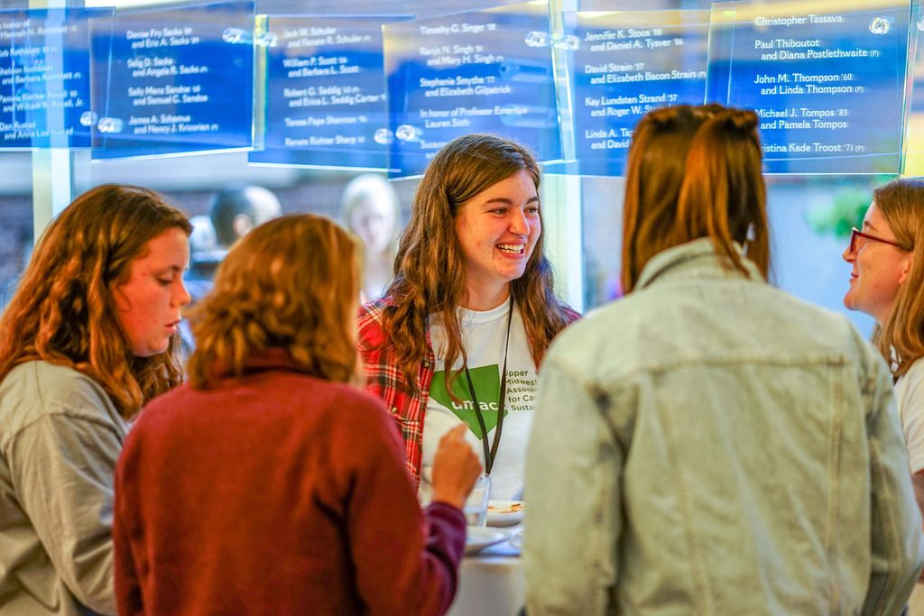 A group of students stand, smile and eat together in a hallway of the Weitz.