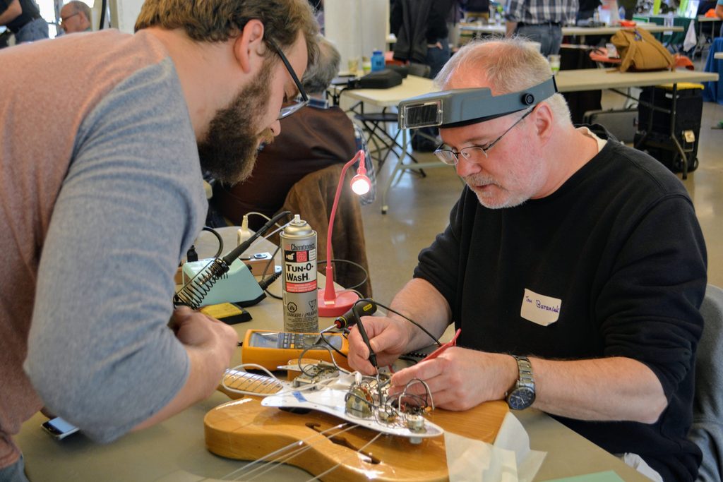 Two people bend over an electric guitar to fix the electronics inside.