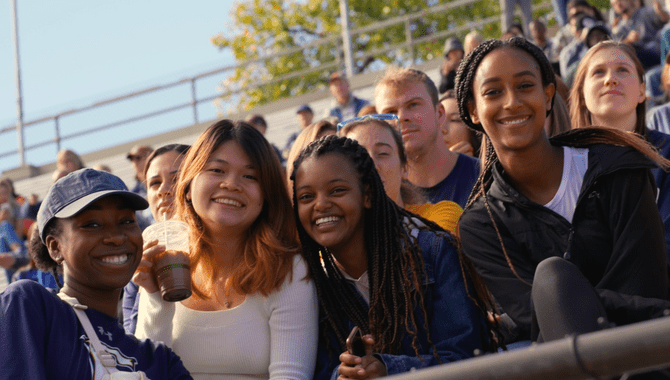 Four students pose and smile together in the stands of a Carleton football game.