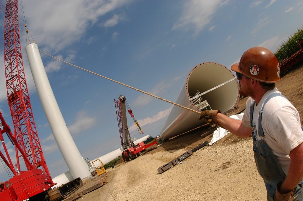 A construction worker holds a long cable attached to a piece of a wind turbine, mid-construction.