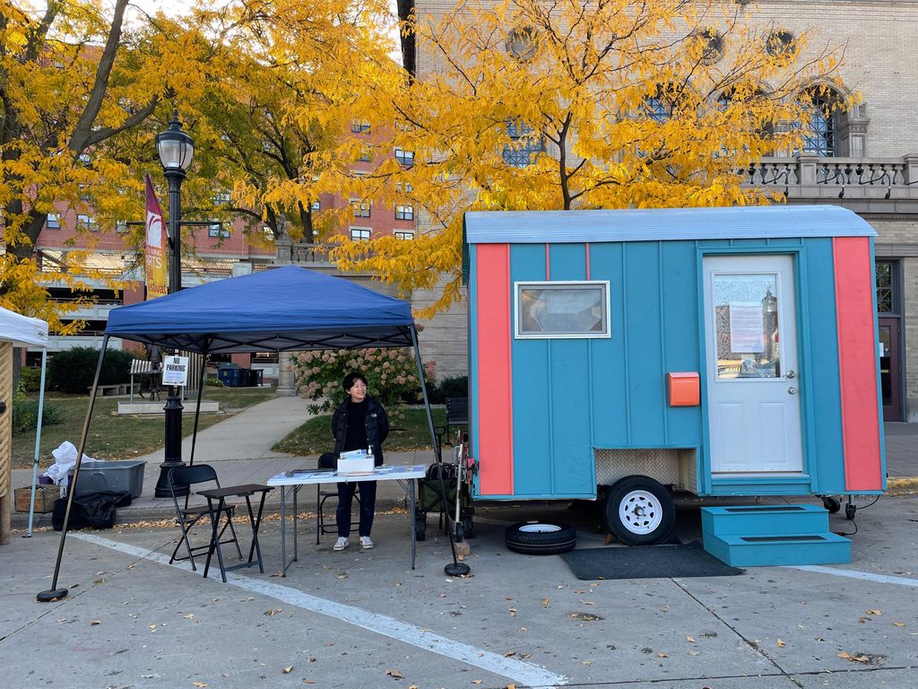 Peixuan Ouyang '18 stands at a table under a tent next to the Wandering House