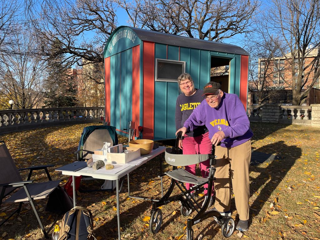 An older person with a walker and a person in a Carleton sweatshirt pose in front of the Wandering House.