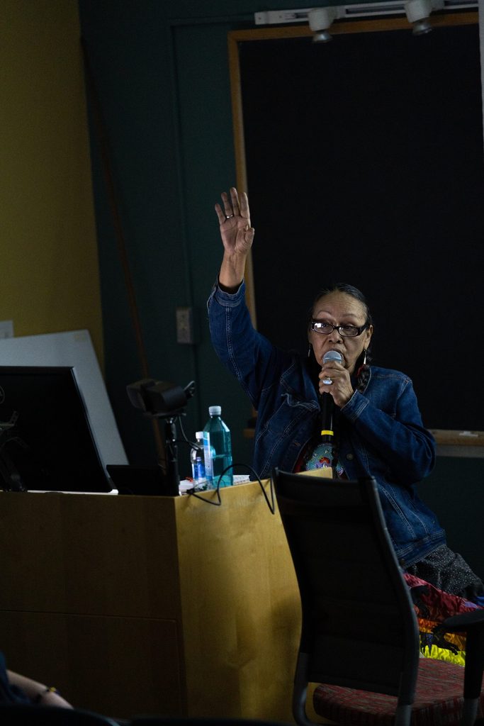 Ida Downwind speaks into a microphone and raises a hand in a dimmed classroom.