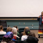 Ida Downwind speaks while holding papers at the front of a classroom while people look on from chairs.