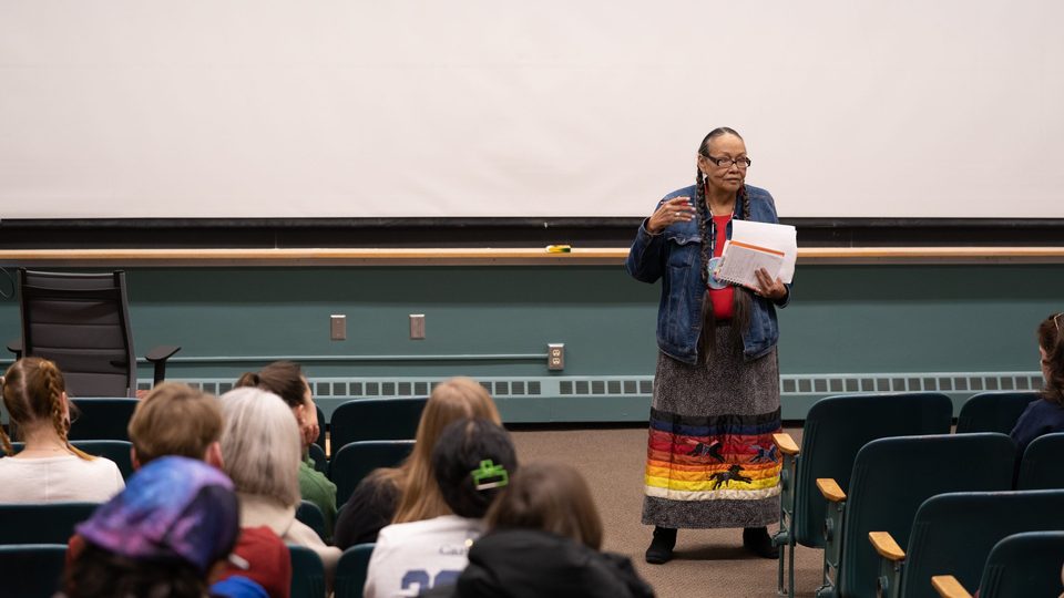Ida Downwind speaks while holding papers at the front of a classroom while people look on from chairs.