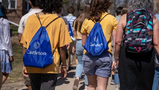 A tour group walks away as the camera focuses on two students wearing yellow shirts and blue Carleton drawstring bags.