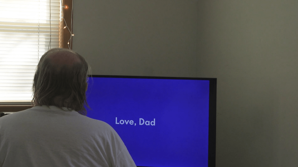 Man sits staring at television screen which reads "Love, Dad"