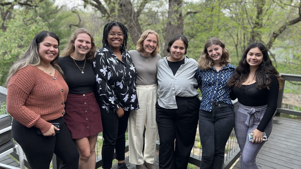 Seven students pose together on a wooden deck outside.