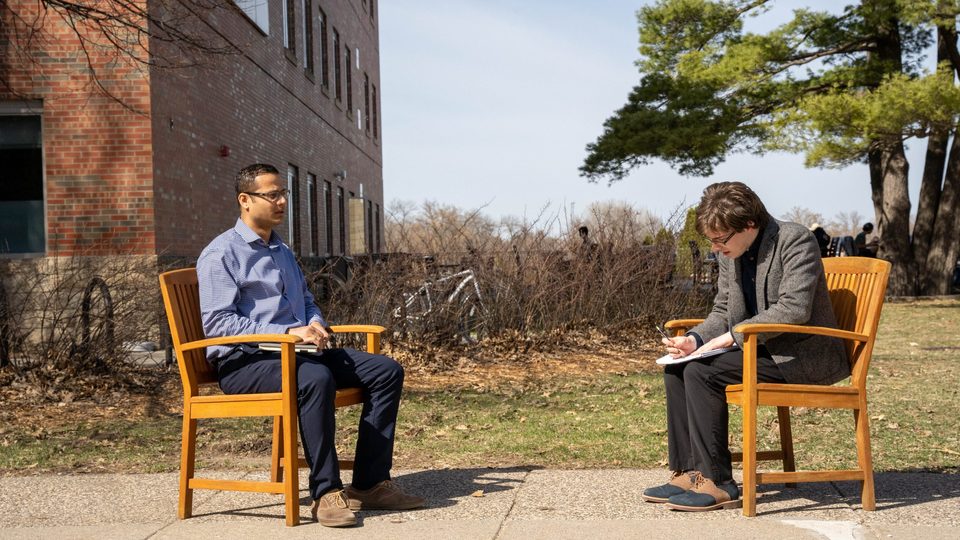 A professor and a student sit opposite each other on two benches, outside in the sun.