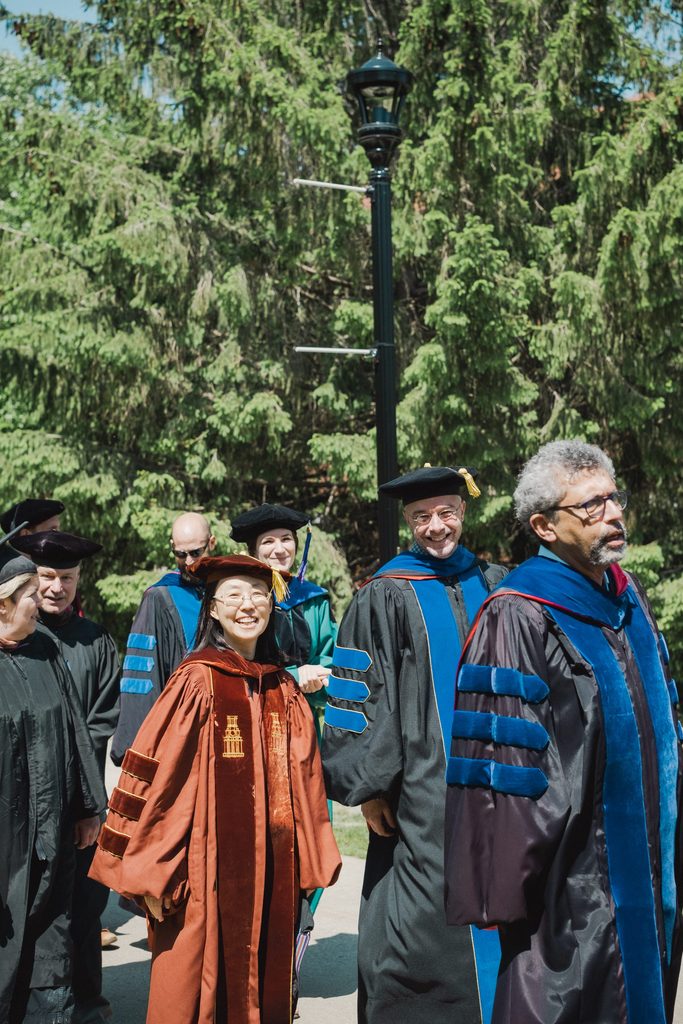 Professors in full regalia smile at the camera as they walk in their processional.