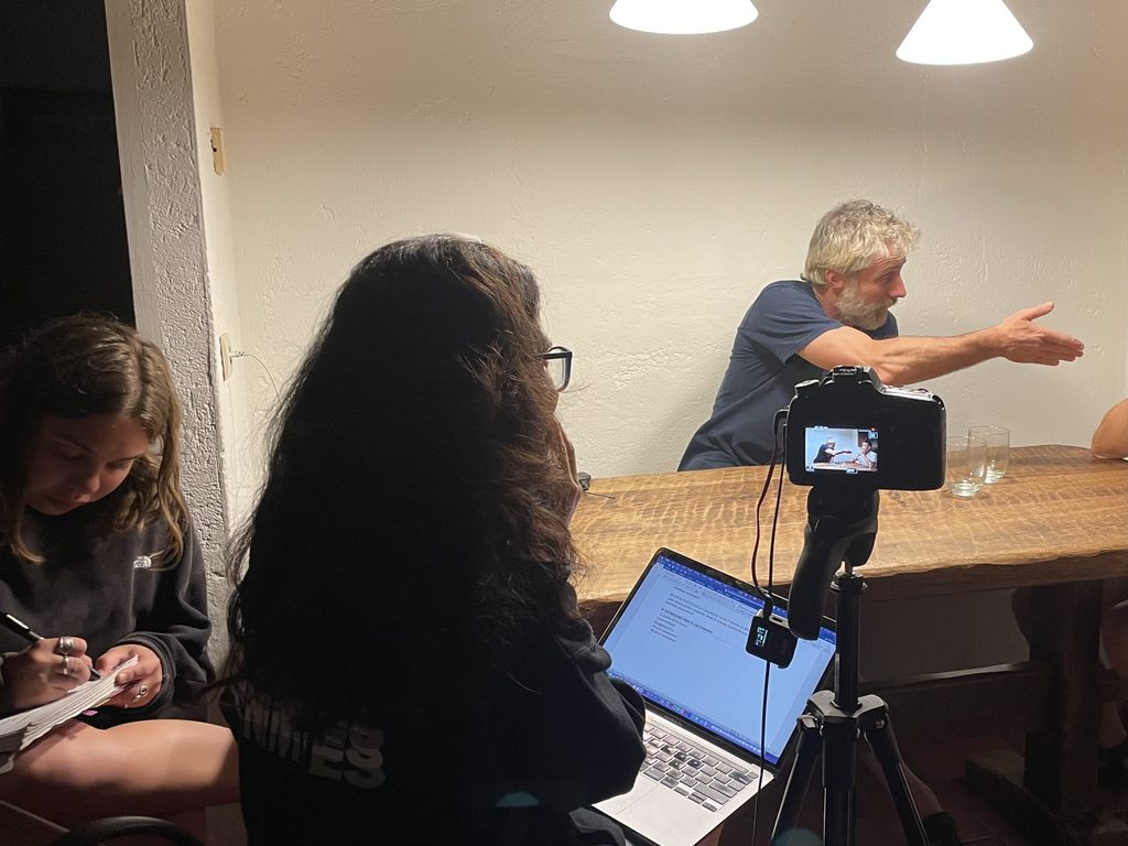 A person gestures to something off camera as a student takes notes and a professor records the person with a video camera and laptop.