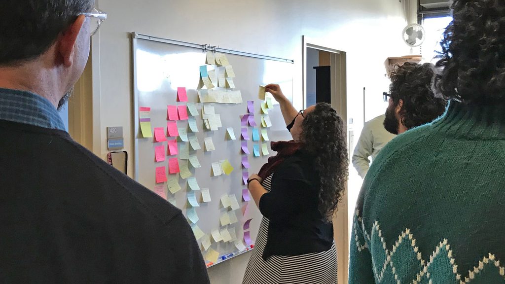A woman adds sticky notes to a whiteboard while several men observe