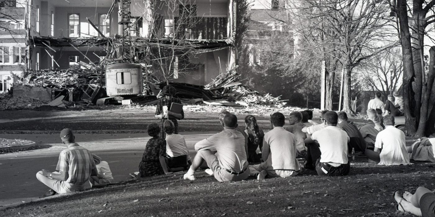 students sit on a hillside watching a building being torn down