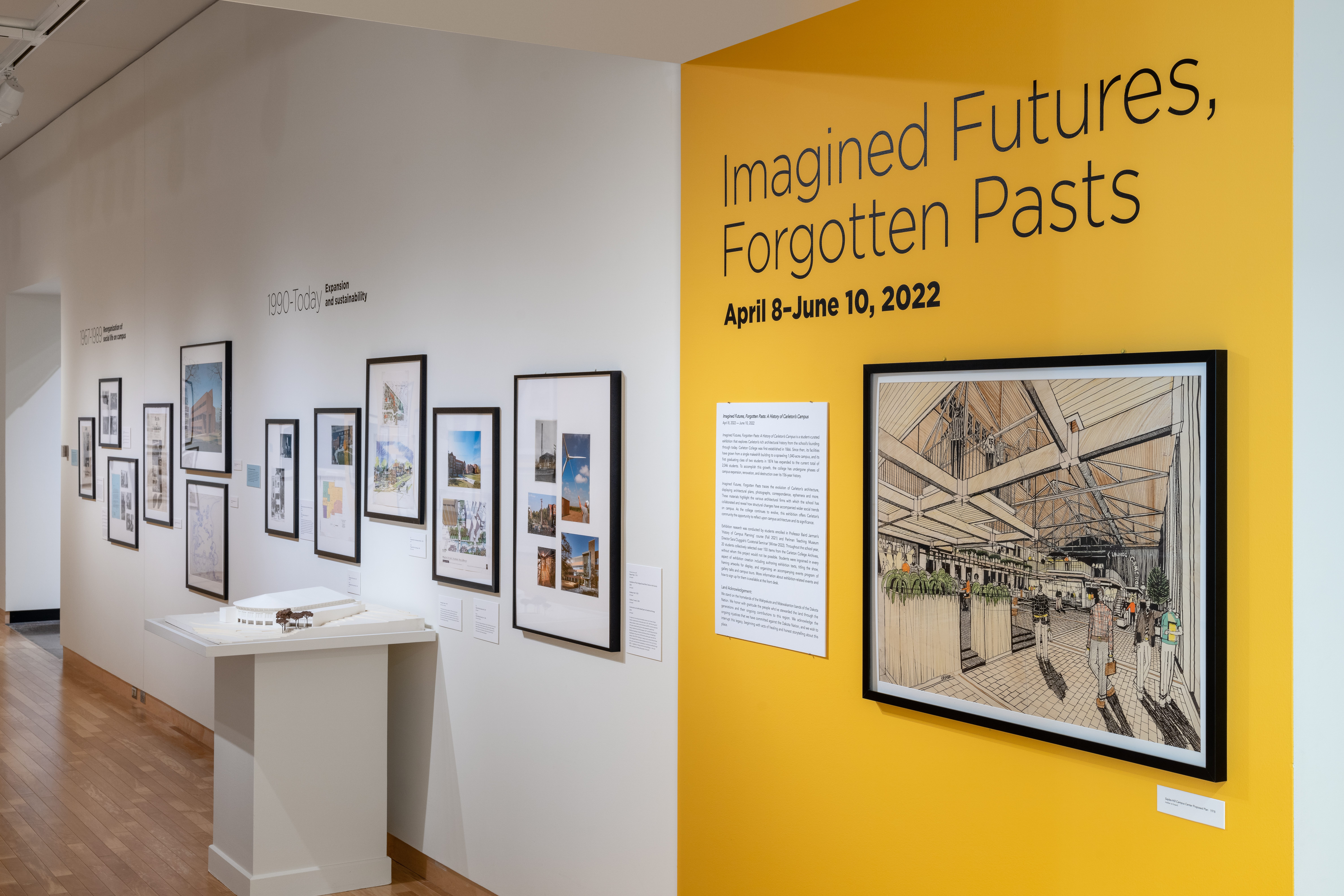 “Imagined Futures, Forgotten Pasts” painted in black text on a yellow wall above a large drawing of the Sayles-Hill Campus Center remodel and next to smaller framed photographs on a white wall