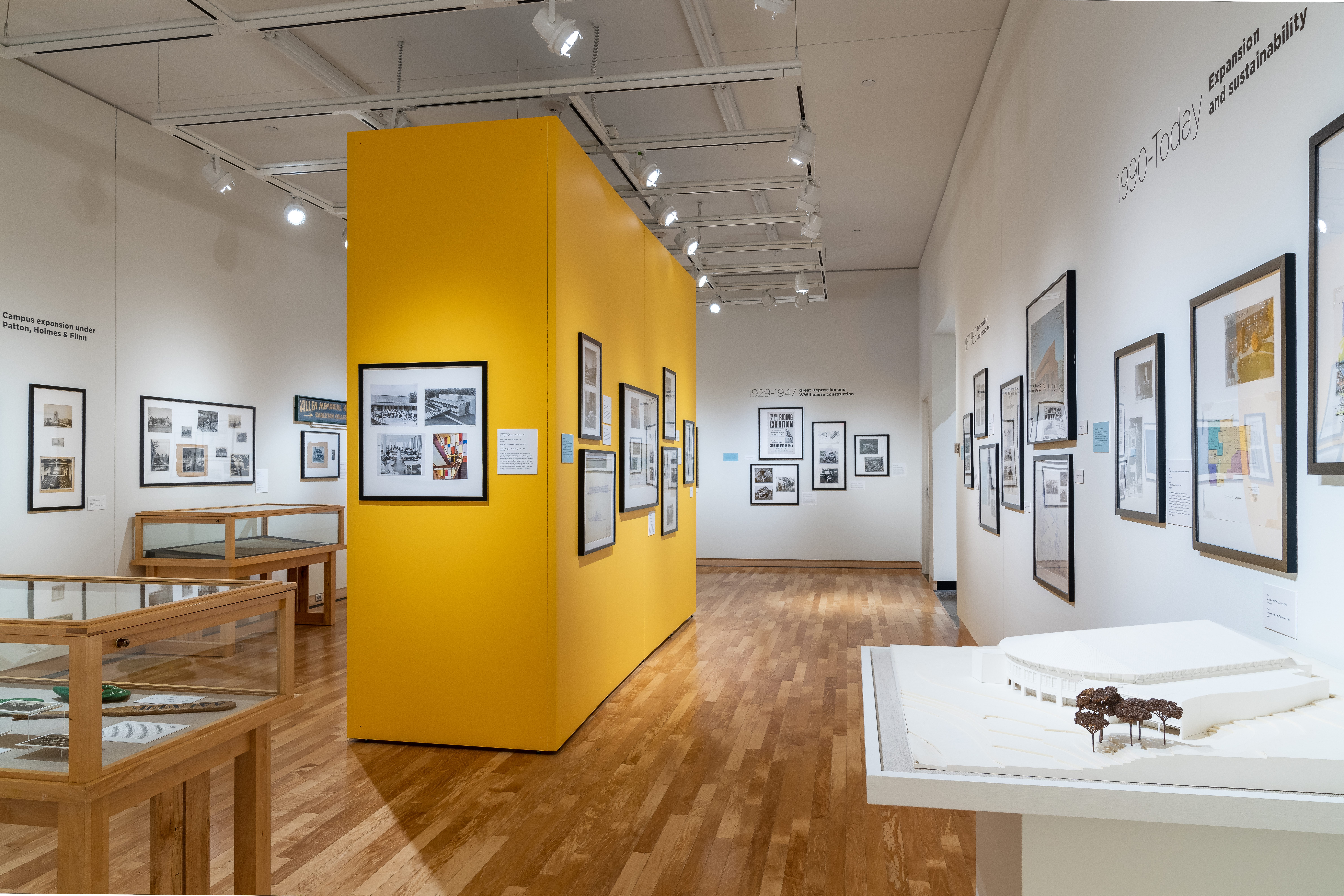 Photo of the yellow wall in the center of the gallery, with framed hanging images and glass display cases on two sides