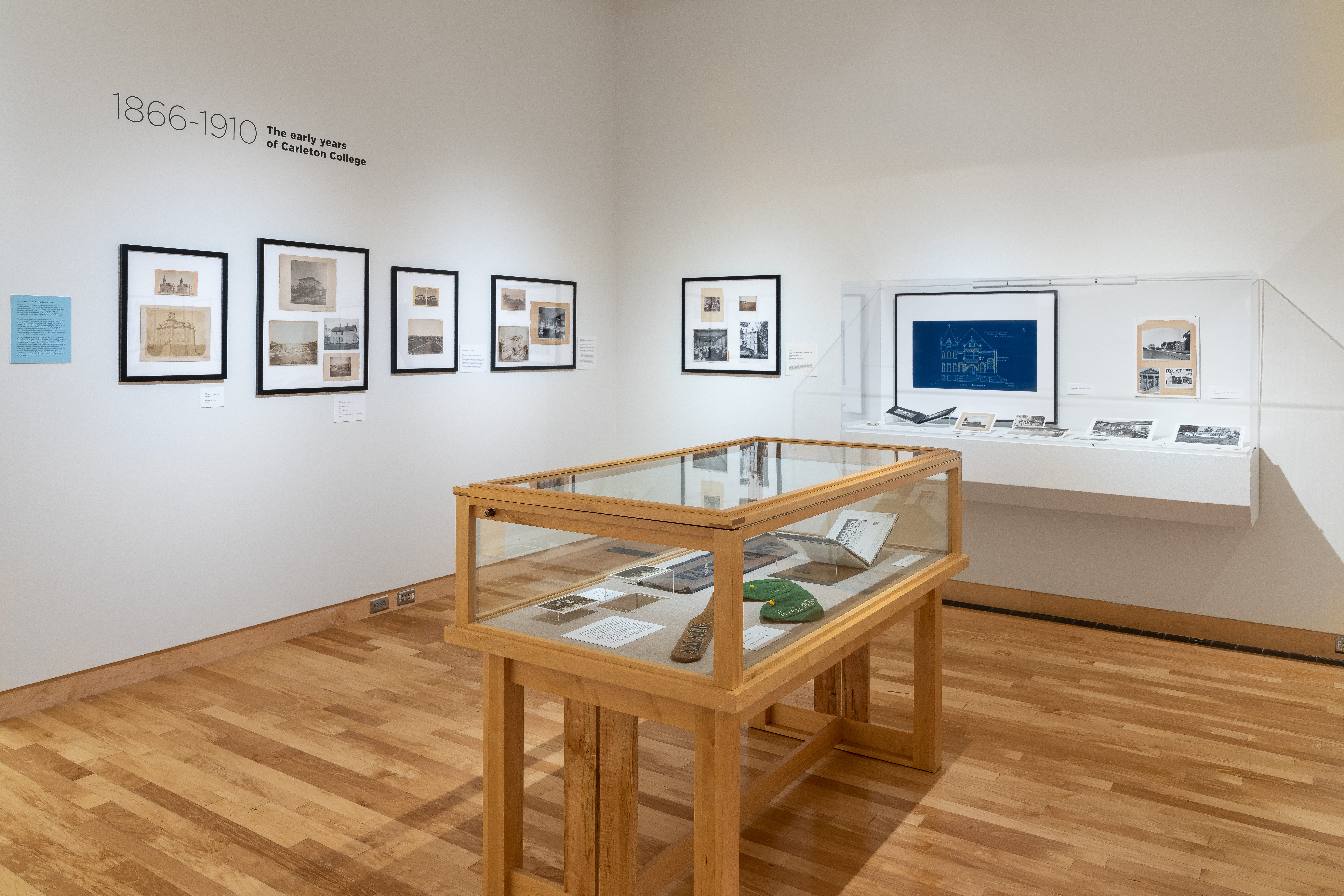 Photograph of a corner of the gallery, with a display case in front of hanging images from 1866-1910
