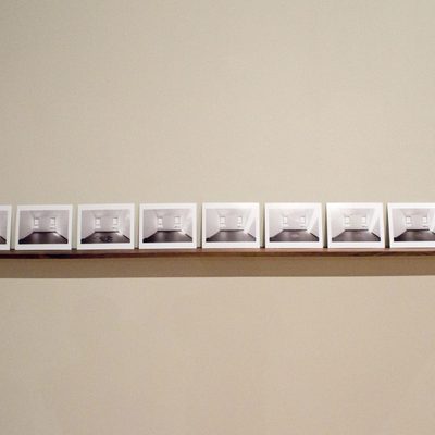 A horizontal line of black and white photos on a wooden shelf all showing the same white room with two parallel, rectangular windows photographed from straight on. In a few of the images it looks like there is something on the gray floor of the room