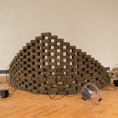 Brown bricks form a triangular wall wrapped in a semicircle. Gaps between the bricks allow you to see between them. A pile of stones sits to the side and speakers sit around the wall.