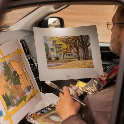 Older man painting a still life of a house with fall leaves while sitting inside his car. The photo is taken through the driver’s window