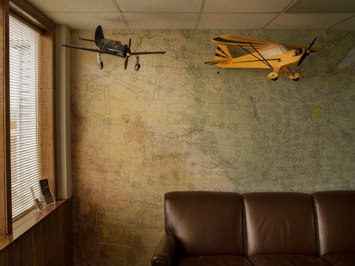Two model airplanes, one black and one yellow, hang from the ceiling in an old building. A faded green and beige map covers the wall behind them