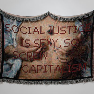 A rectangular cotton cloth with fringe that shows a pixelated image of the exposed breasts of a woman. Red text over the work reads “Social Justice is Sexy, So Screw Capitalism.