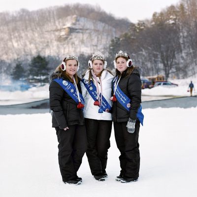 An outdoor portrait of three young women wearing snowsuits and matching tiaras, sashes, and earmuffs