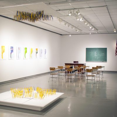 Artistic installation of a classroom. Wooden desks are aligned in front of a green chalkboard. A line of school desktop art installations are along the wall and a white platform on the floor has yellow pencils sticking from it vertically.