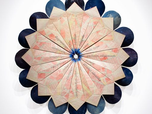 Large artistic paper sculpture shaped like a circular flower with dark blue edges and a light pink center constructed from paper with a faint pattern and words printed on it.