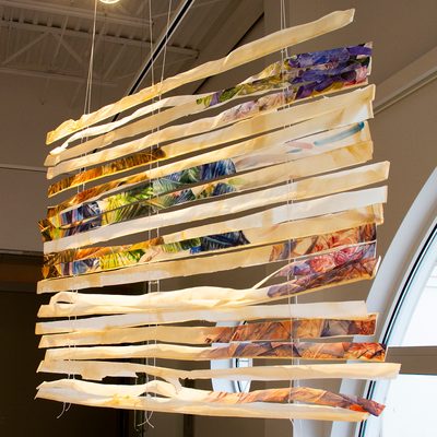 A series of horizontal strips of paper which are attached to the ceiling by a string and hang together like blinds for a window. The strips of paper are painted with watercolor images of vibrantly colored flowers and leaves. The images turn into more abstract brushstrokes, and some of the strips are entirely covered by a light brown wash.