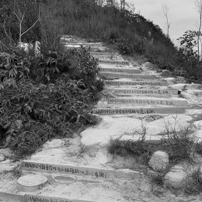 [Black and white photo of a trail towards the mountain; Chinese sentences painted on the steps indicating people who maintained the path.