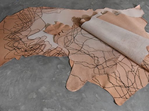 A photograph of a piece of vegetable tanned leather laid on a gray concrete ground. The leather is folded in some places, and is darker around the edges. It is covered with wavy scrawls made with permanent marker