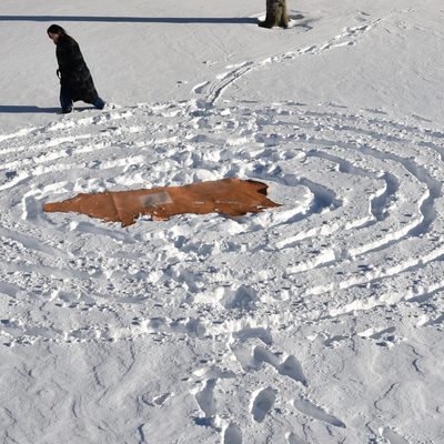A photograph of a piece of beige leather placed onto the ground which is covered by a layer of snow. Around the leather are deep five rings in the snow that were made by footsteps. Behind this scene there is a figure standing knee-deep in the snow.