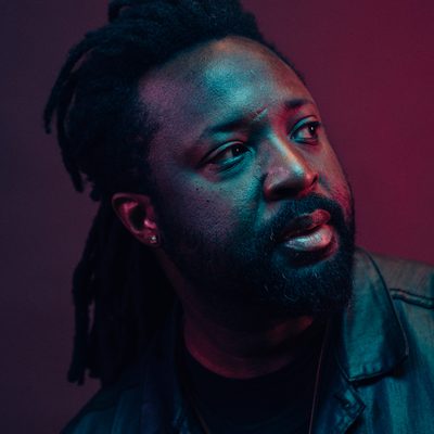 A portrait photograph of author Marlon James, lit with vibrantly colored lights. Marlon looks to the right side of the image, away from the viewer. Purple, teal and magenta light create colorful gradients on his skin. His black leather jacket picks up the teal light. The background is a plain gradient from purple to magenta.