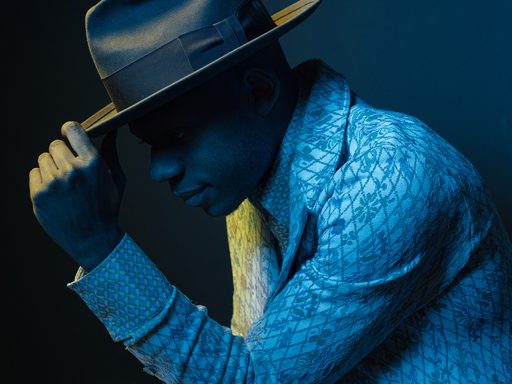 A portrait photograph of musician Ondara in profile facing to the left. His left elbow rests on his left knee, with hand touching the tip of a wide brimmed hat. He wears a light colored suit with a diamond and floral pattern. The image overall is dark blue, skin and suit all taking the blue tone of light, with yellow/green light outlining and highlighting the front of his face and the hand touching the hat. The background is solid darker blue