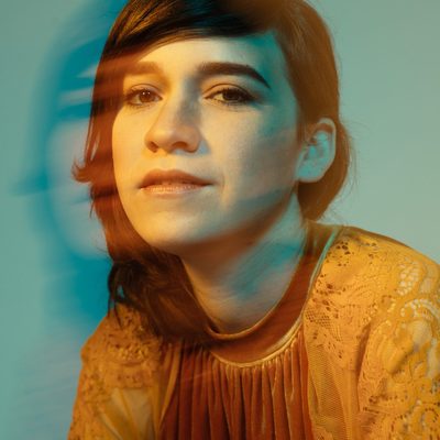 A portrait photograph of musician Reina Del Cid, shoulders and head, looking towards the camera. A gold/orange top with lace sleeve and velvet chest. Orange light illuminates the left half of her face. Teal light blends with the orange light on the right, leading to darker teal shadows on the right side of her face. A faint ghost image of her face streaks to the left from a motion effect, showing a faint version of her face again slightly offset to the left. The background is a plain teal, with some gradation