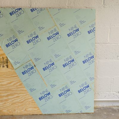 A piece of plywood that is decorated with light blue posters that read “Nine Below Zero” in darker blue lettering. The posters are applied to the board diagonally, and the bottom left corner is left exposed
