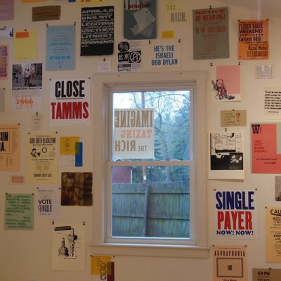 A series of multicolored printed posters displayed around a window. Some posters have a design, others have phrases like “Single Payer, Now! Now!” and “We are not born equal. We become equal in struggle.” Some reference political issues, other reference musicians, all feature graphic design